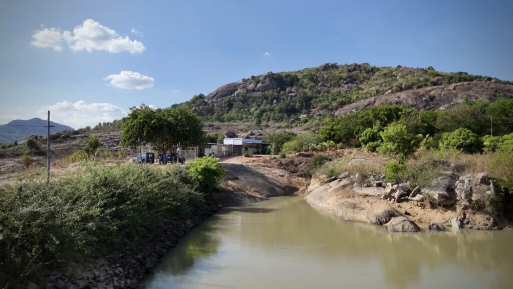 A small river flowing through a rural landscape with green trees, rocky terrain, and hilly mountains in the background on a sunny day with blue sky and clouds.