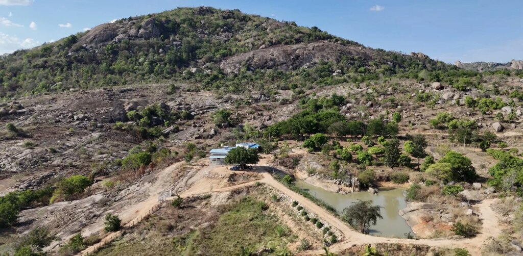A mountain landscape with a small house near a winding dirt road and pond, sparse vegetation, and a rocky hill in the background under a partly cloudy sky.