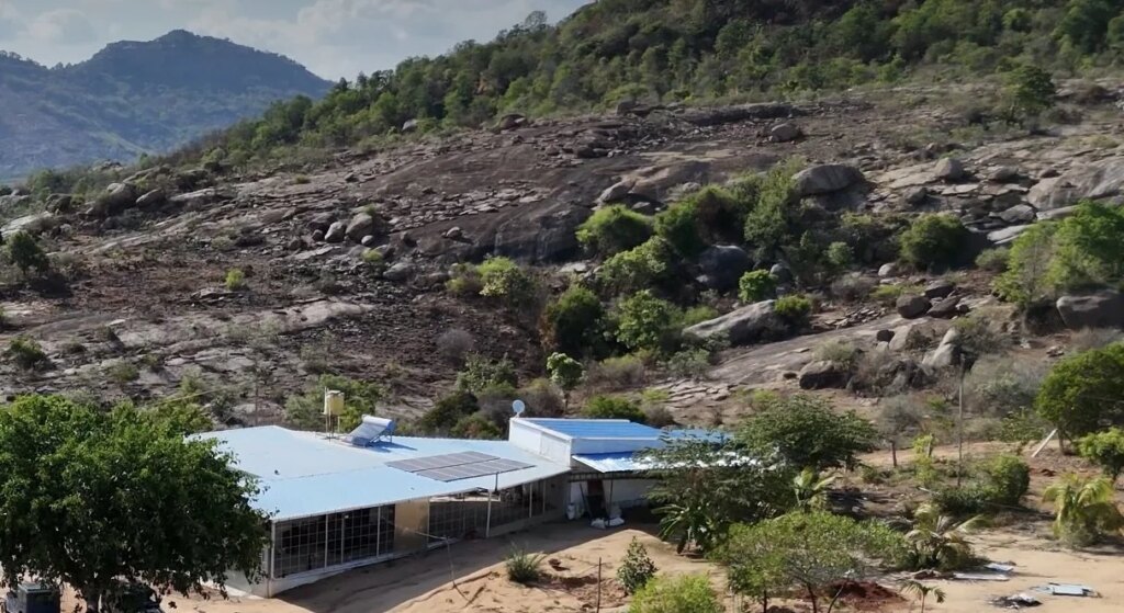 A house with solar panels on its roof in a rural area surrounded by trees, with rocky hills and mountains in the background.